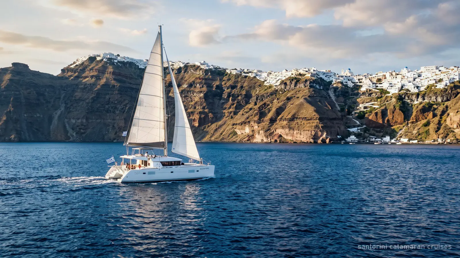 A white sailing catamaran crossing the Santorini caldera below the cliff villages of Fira and Imerovigli